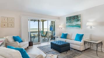 A living room with a sofa, ottoman, and chairs, a balcony with outdoor furniture, and a painting on the wall.
