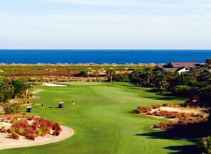 A golf course with a view of the ocean, surrounded by greenery and a few buildings.