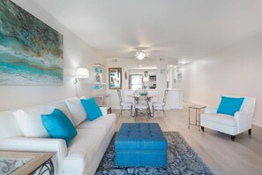 A living room with a sofa, ottoman, and armchairs, a dining area with a table and chairs, and a kitchen in the background.
