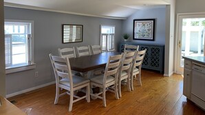 Comedor con mesa de madera y seis sillas, pared gris con dos ventanas, y zona de cocina con armarios y encimera.