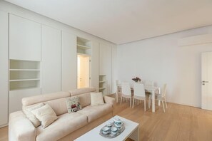A modern living room with a beige sofa, white dining table, and chairs. There is a white cabinet with shelves and a door leading to another room.