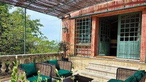 A covered outdoor seating area with wicker furniture and green cushions, a stone balustrade, and a view of a building with large windows and a green door. - Villa Ocre Menerbes Panoramic view Swimming pool Garden Terraces (MENERBES)