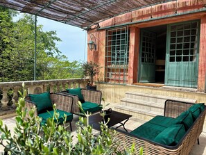 A covered outdoor seating area with wicker furniture and green cushions, a stone balustrade, and a view of a building with large windows and a green door. - Villa Ocre Menerbes Panoramic view Swimming pool Garden Terraces (MENERBES)