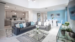 Un salon moderne avec un canapé gris, une table basse en verre et un coin repas agrémenté d’un mur bleu et de chaises blanches.