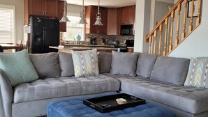 Ein Wohnzimmer mit einem grauen Ecksofa, einem blauen Hocker und einem weißen, flauschigen Teppich.