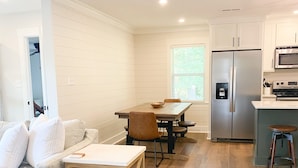 A modern kitchen with white cabinetry, a stainless steel refrigerator, and a microwave. There is a dining area with a wooden table and chairs, and a living room with a sofa and a coffee table.