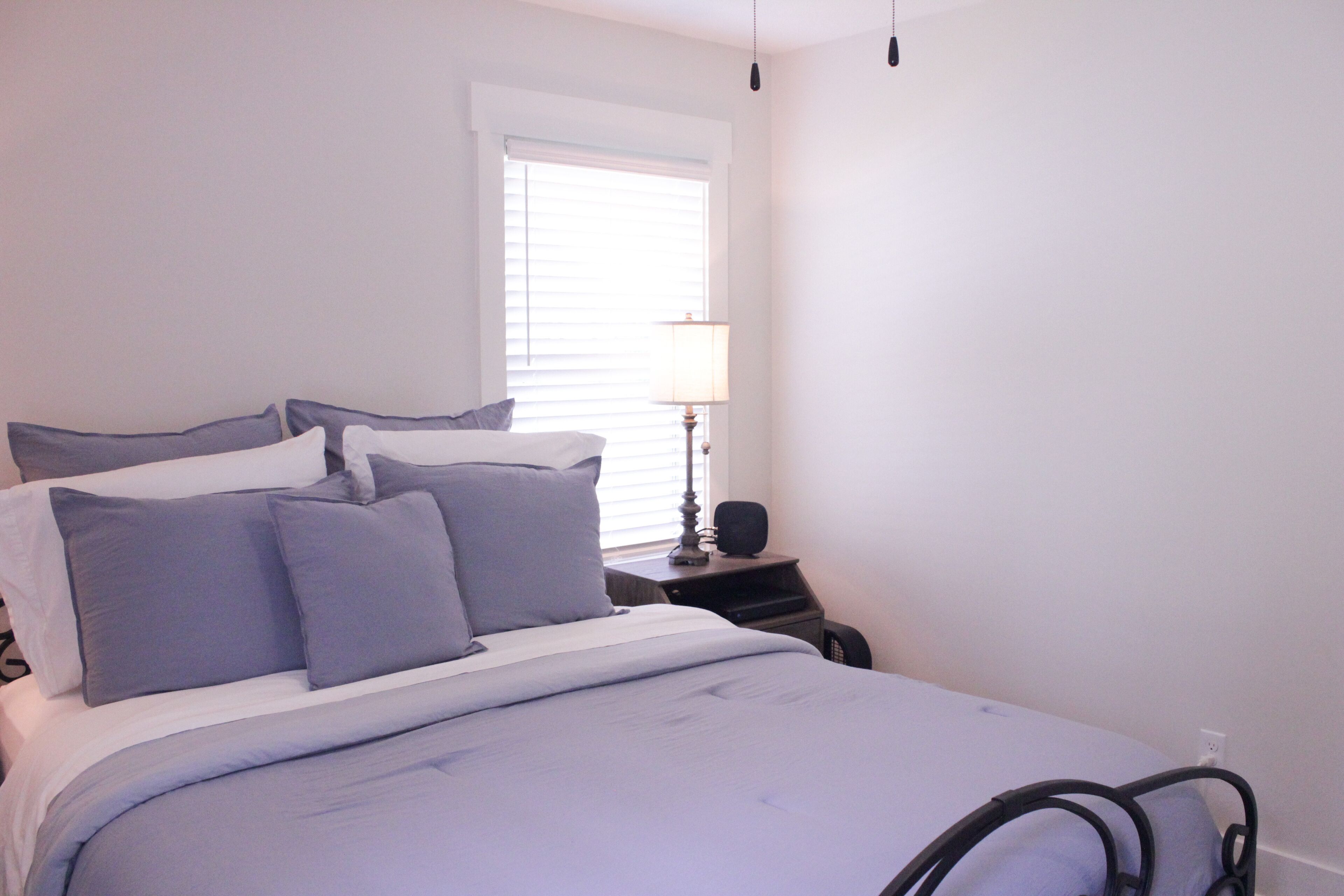 A neatly made bed with a white and blue color scheme, a bedside table with a lamp, and a window with blinds.