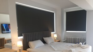 Ein modernes Hotelzimmer mit einem großen Bett, zwei Nachttischen mit Lampen, einem Wand-Fernseher und einem Fenster mit Jalousien.