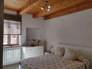 A bedroom with a wooden ceiling, a large bed with floral bedding, a dresser, a mirror, and a window with blinds.
