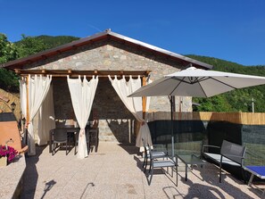 A stone-roofed outdoor seating area with tables and chairs, an umbrella, and a view of a forested hill.