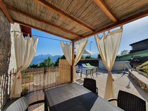 A covered outdoor seating area with a table and chairs, a view of mountains, and a stone wall.