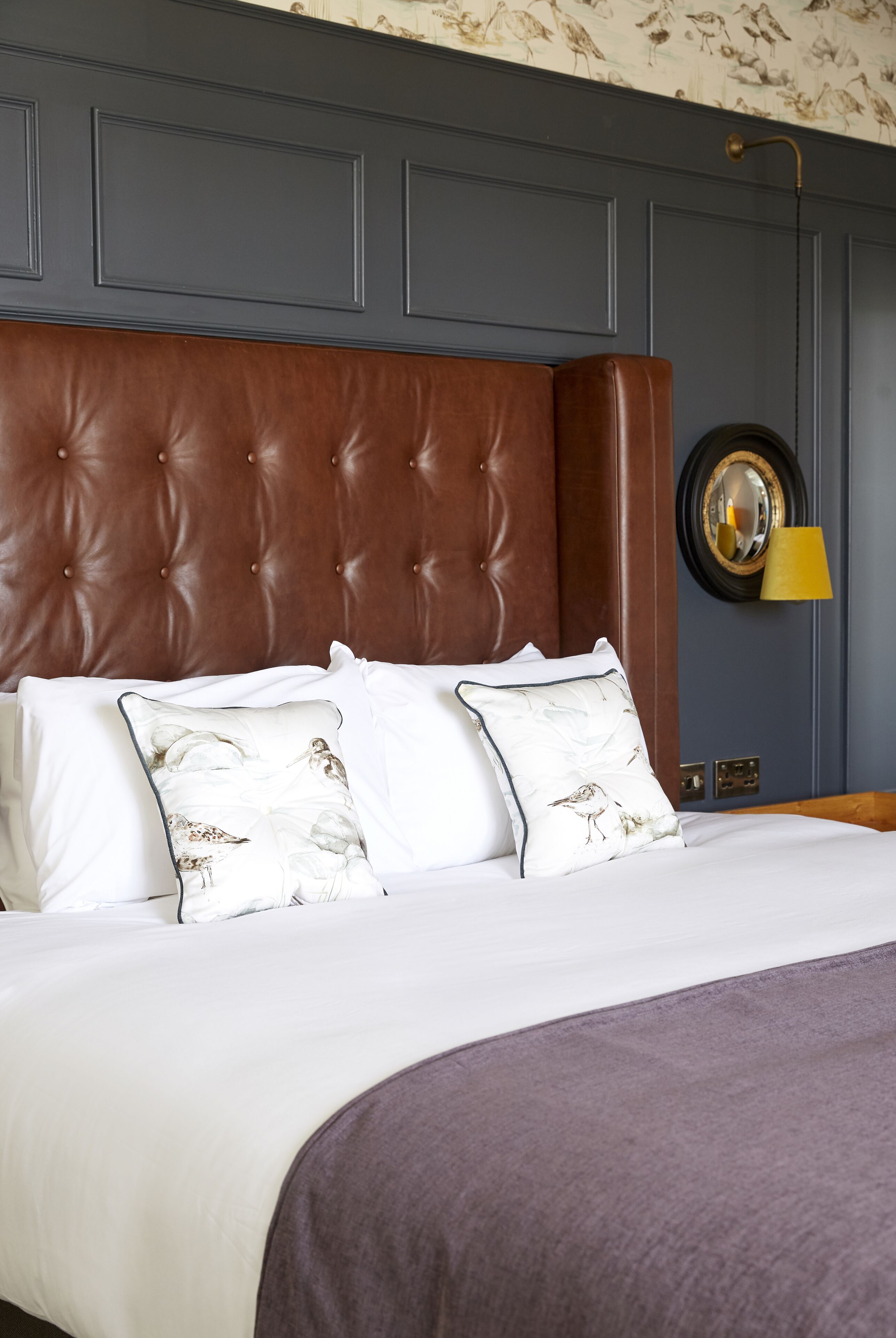 A neatly made bed with a brown headboard, white bedding, and two decorative pillows with a bird pattern.