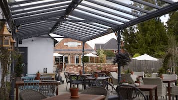 Ein Terrassenbereich im Freien mit Tischen und Stühlen, ein überdachter Sitzbereich und im Hintergrund ein Gebäude mit der Aufschrift 'Coach House Bar'.