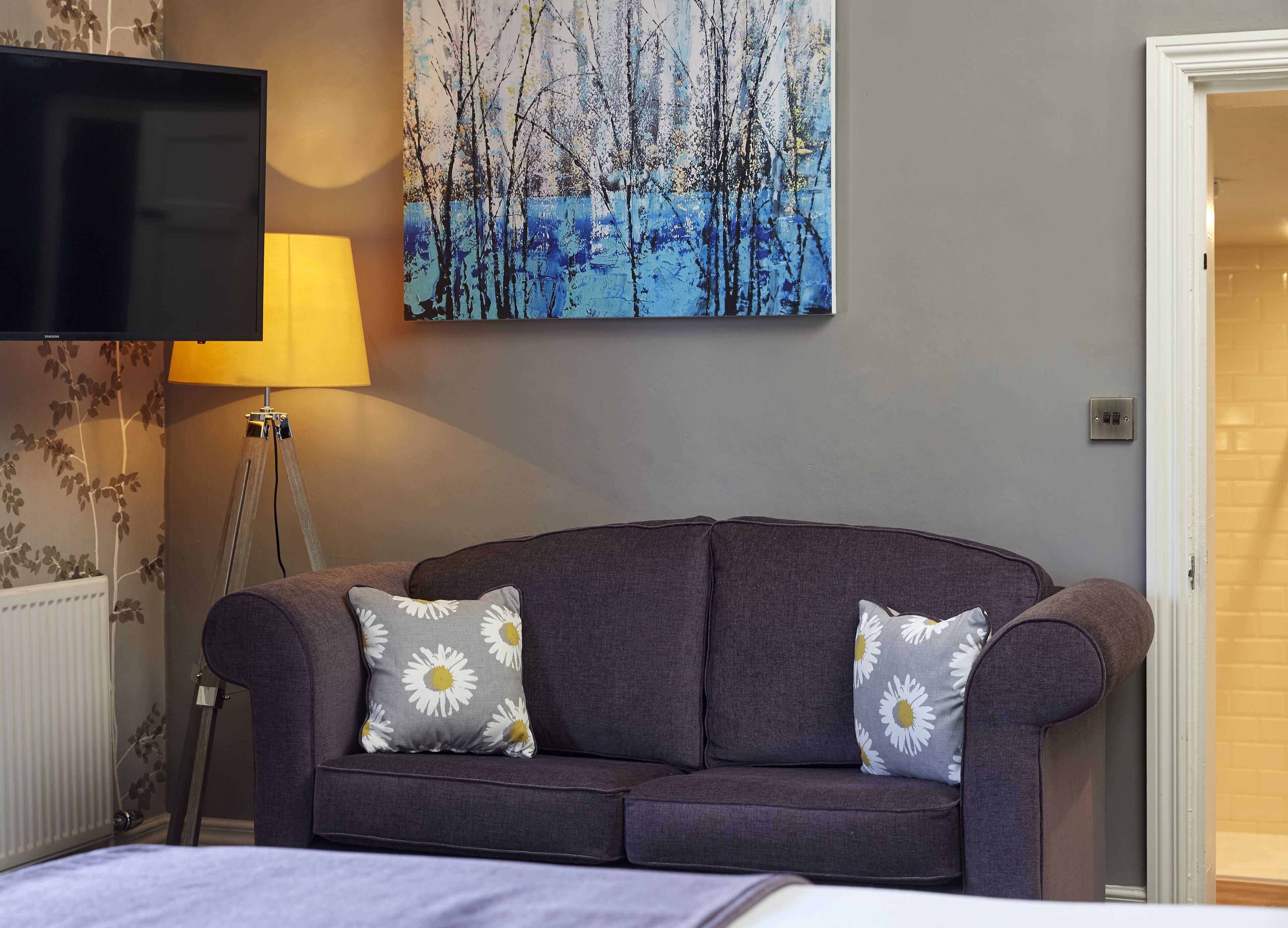 Un salon moderne avec un canapé gris, deux coussins assortis, un téléviseur à écran plat, une lampe sur pied et un tableau représentant des arbres.