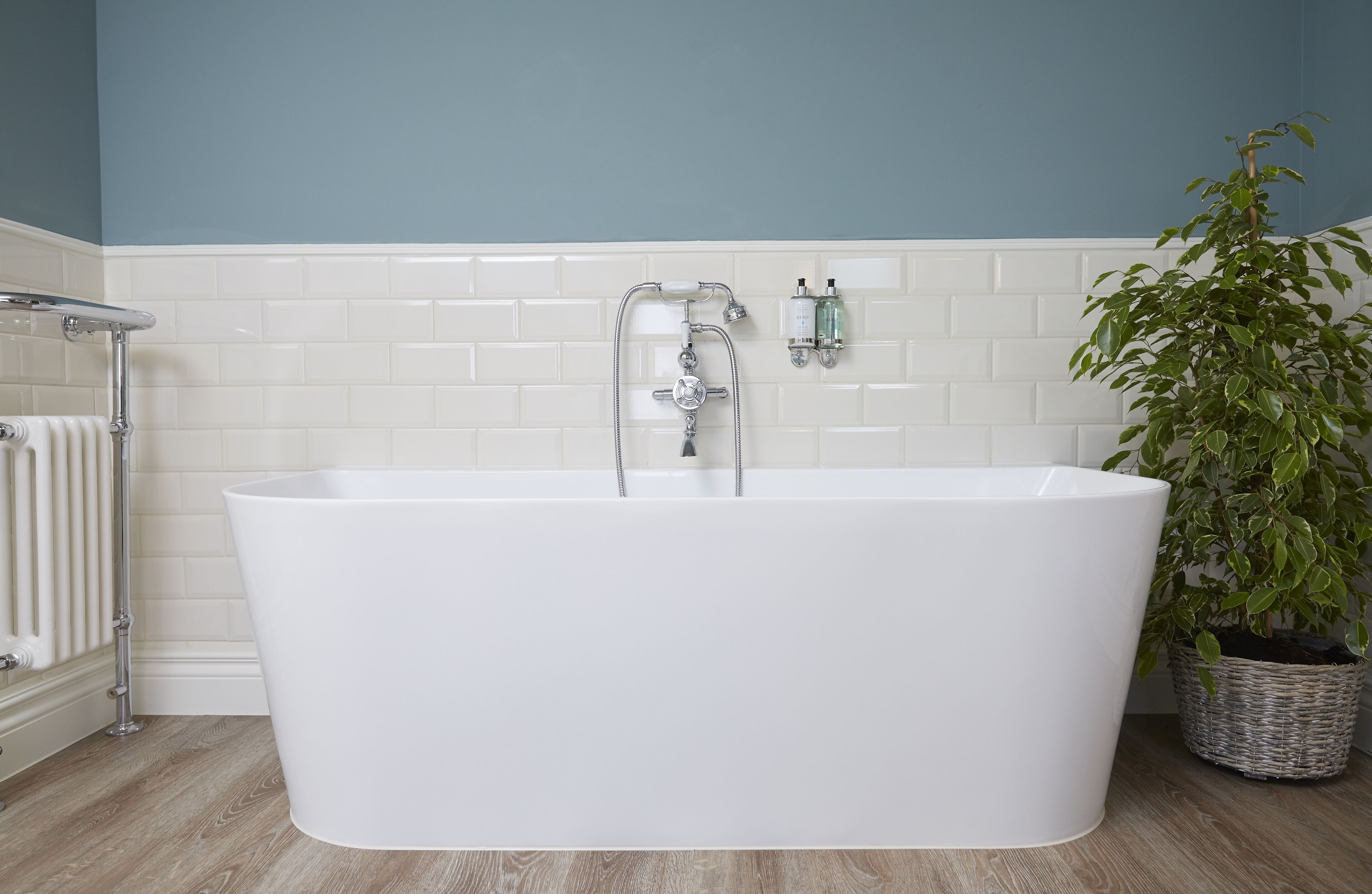 Une baignoire blanche, autoportante, dans une salle de bain aux murs recouverts de carreaux de céramique blancs, avec un radiateur et une plante en pot.