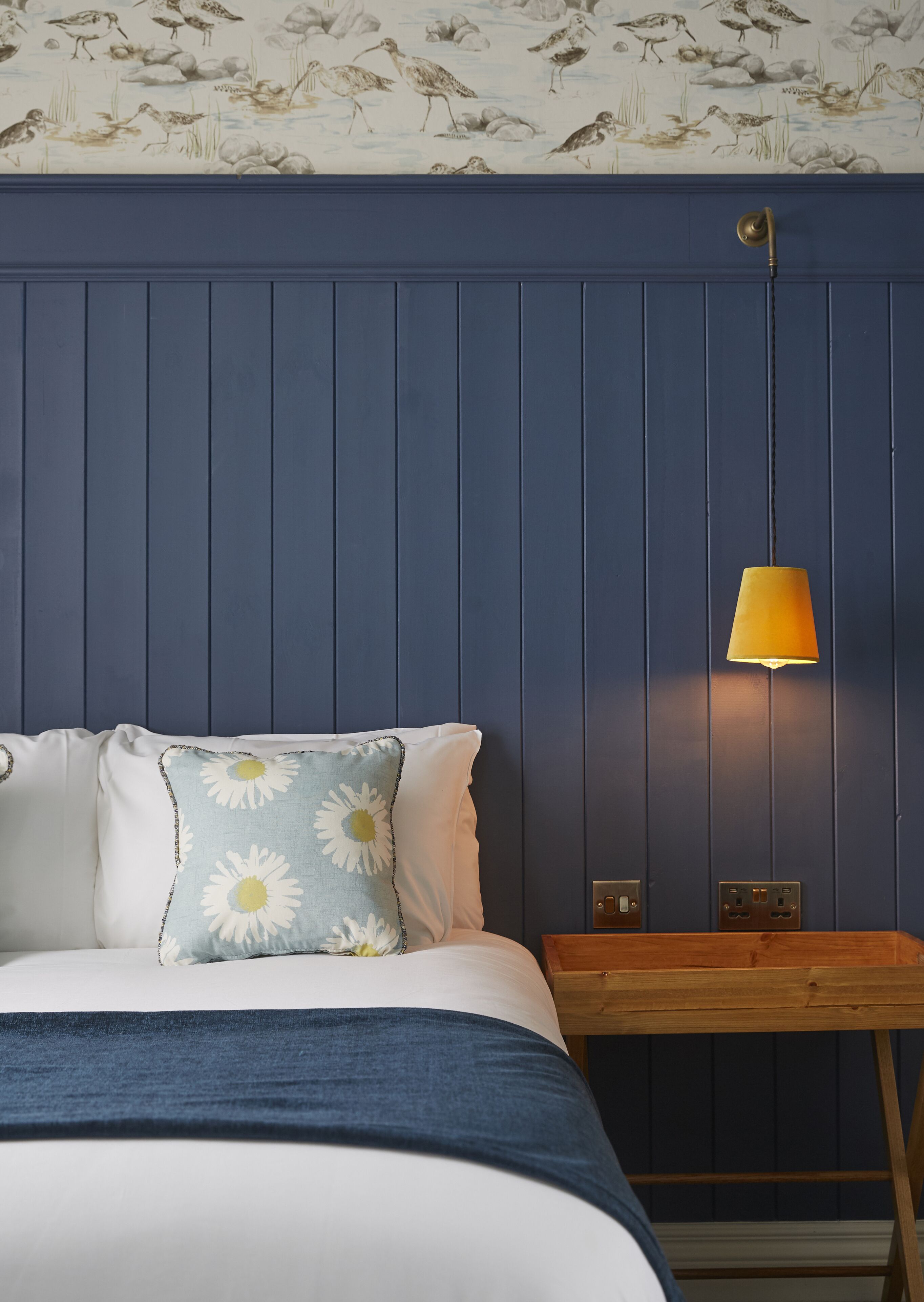 Un dormitorio con cama, almohadas, mesita de noche, lámpara y pared con estampado de pájaros.