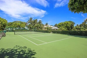 A tennis court with a net, surrounded by greenery and a building in the background. - Captiva Tarpon (Captiva)