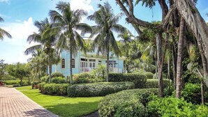 Un edificio azul claro con un balcón, rodeado de exuberante jardinería tropical con palmeras y setos.