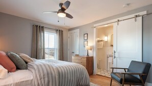 A bedroom with a bed, a chair, a ceiling fan, a window with curtains, and a bathroom visible through an open door. - Pharmacy Apartment | Luxury Monthly Stay Downtown (Charlottesville)