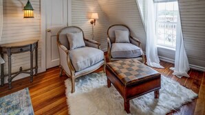 Une pièce mansardée chaleureuse, avec un plancher en bois, deux fauteuils rembourrés, un pouf à carreaux, une petite table et une lampe.