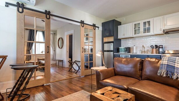 A modern living room with a brown leather sofa, a wooden coffee table, and a sliding barn door leading to a kitchen. - Southern Luxury Apartment | Downtown Charlottesville | 30 Night Minimum (Charlottesville)