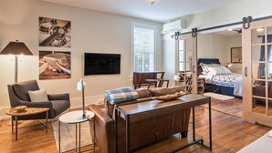 A modern living room with a sofa, coffee table, and a TV. There is a ceiling fan, a window with blinds, and a door leading to another room. - Southern Luxury Apartment | Downtown Charlottesville | 30 Night Minimum (Charlottesville)