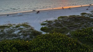 Een strand met een ondergaande zon aan de horizon, mensen die wandelen en ontspannen op het zand, en wat begroeiing op de voorgrond.