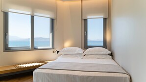 Een moderne slaapkamer met een groot bed, twee ramen met zonwering en uitzicht op zee.