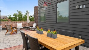 Eine Terrasse im Freien mit einem Holztisch und Stühlen, einer Ziegelwand und einem großen Fenster.