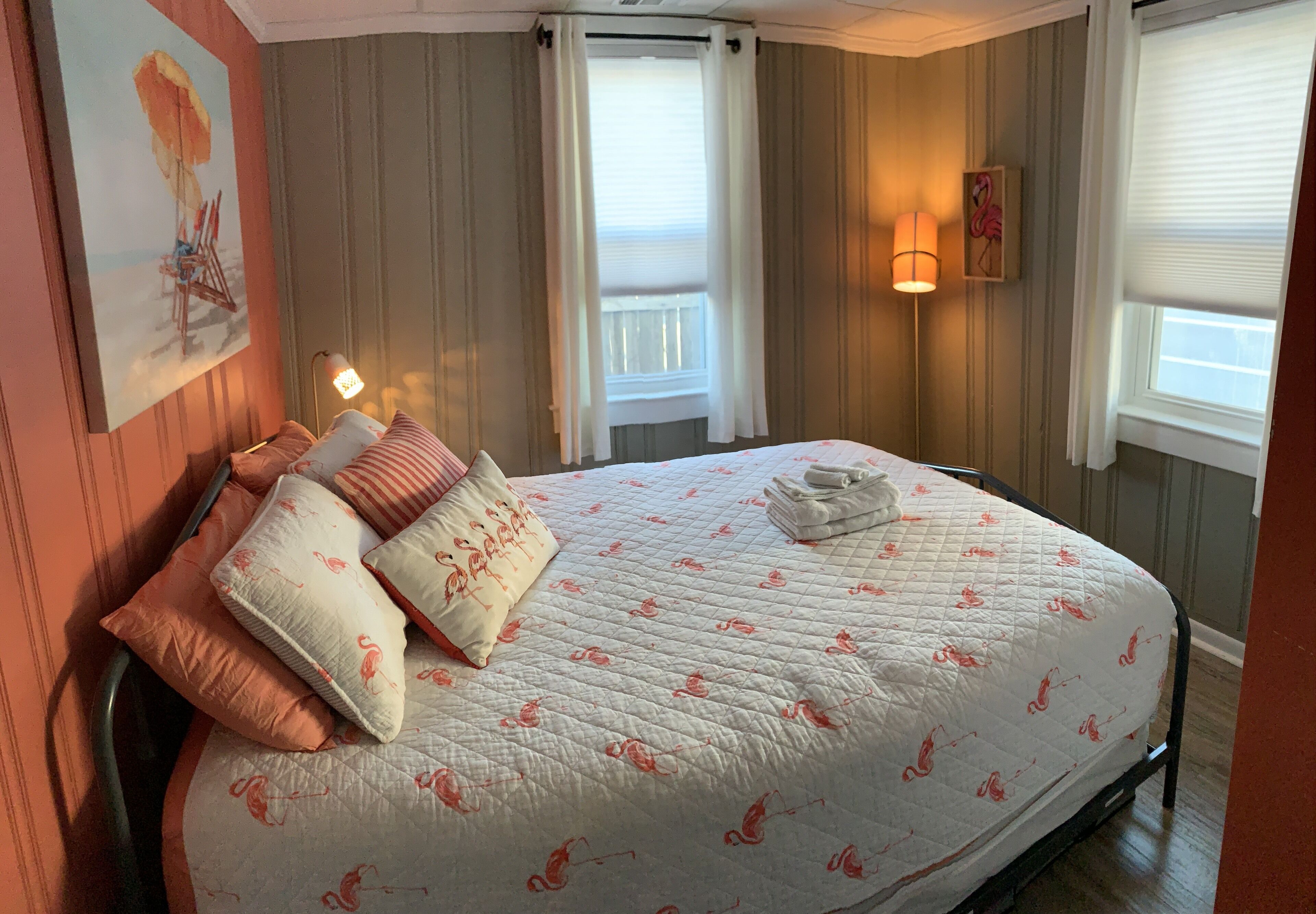 A neatly made bed with a floral quilt, a painting of a beach scene, and a wall-mounted lamp.