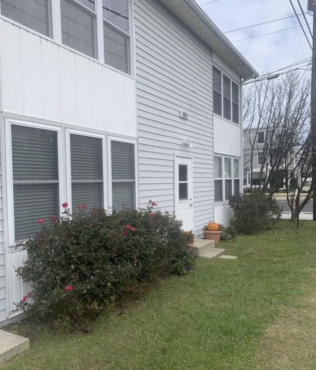 A two-story residential building with a white exterior, a front door, and a small front yard with a bush and a pumpkin planter.