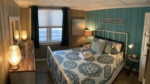Een slaapkamer met een bed, nachtlampjes, een spiegel, een raam met gordijnen en een muur met een 'lets be mermaids' bord.
