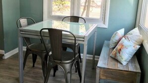 Een eetgedeelte met een glazen tafel en metalen stoelen, een bank met kussens, en een raam met zonwering.