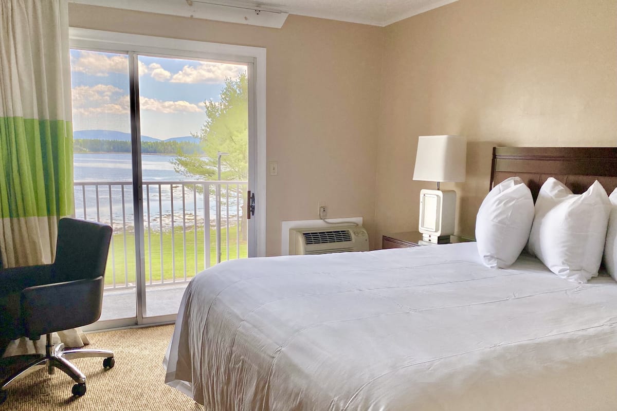 A hotel room with a large bed, a chair, a lamp, and a view of a lake through a sliding door.