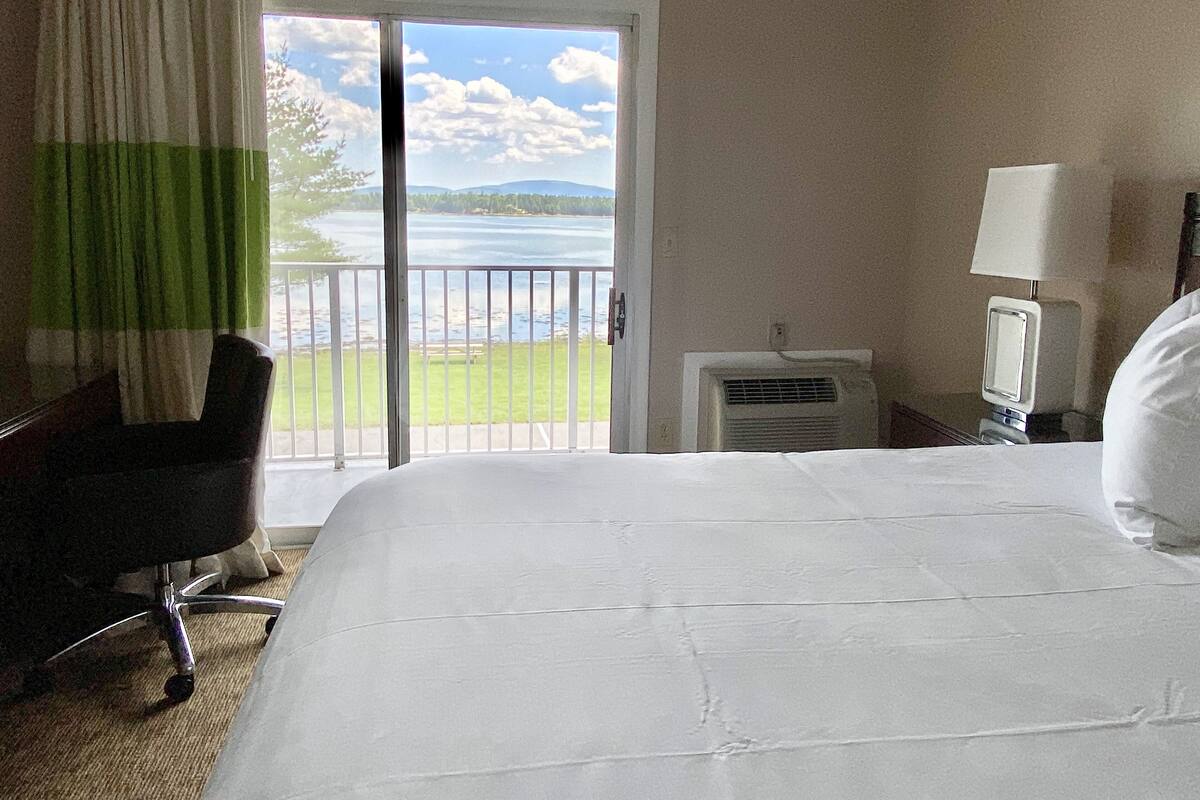 A hotel room with a bed, a chair, a TV, and a view of a lake through a sliding door.