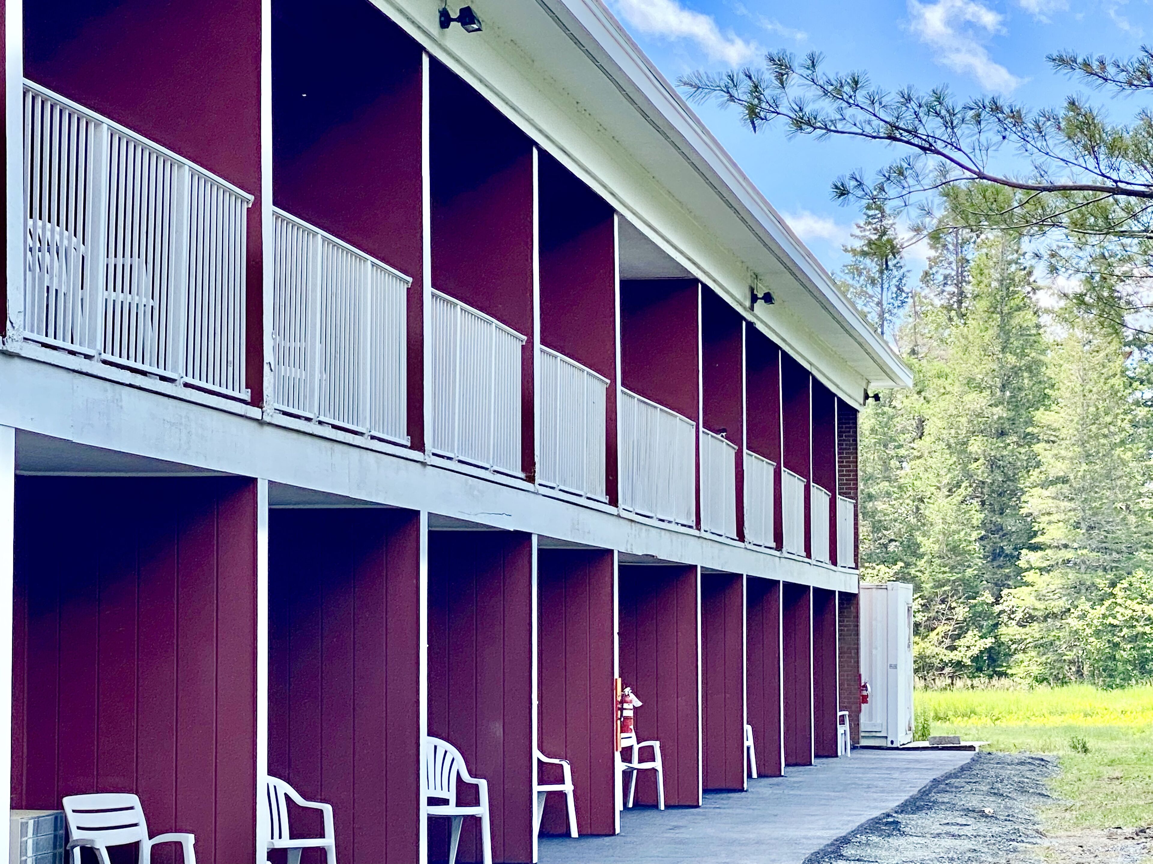 A row of red and white rooms with white railings, a covered outdoor area, and a view of trees and sky.