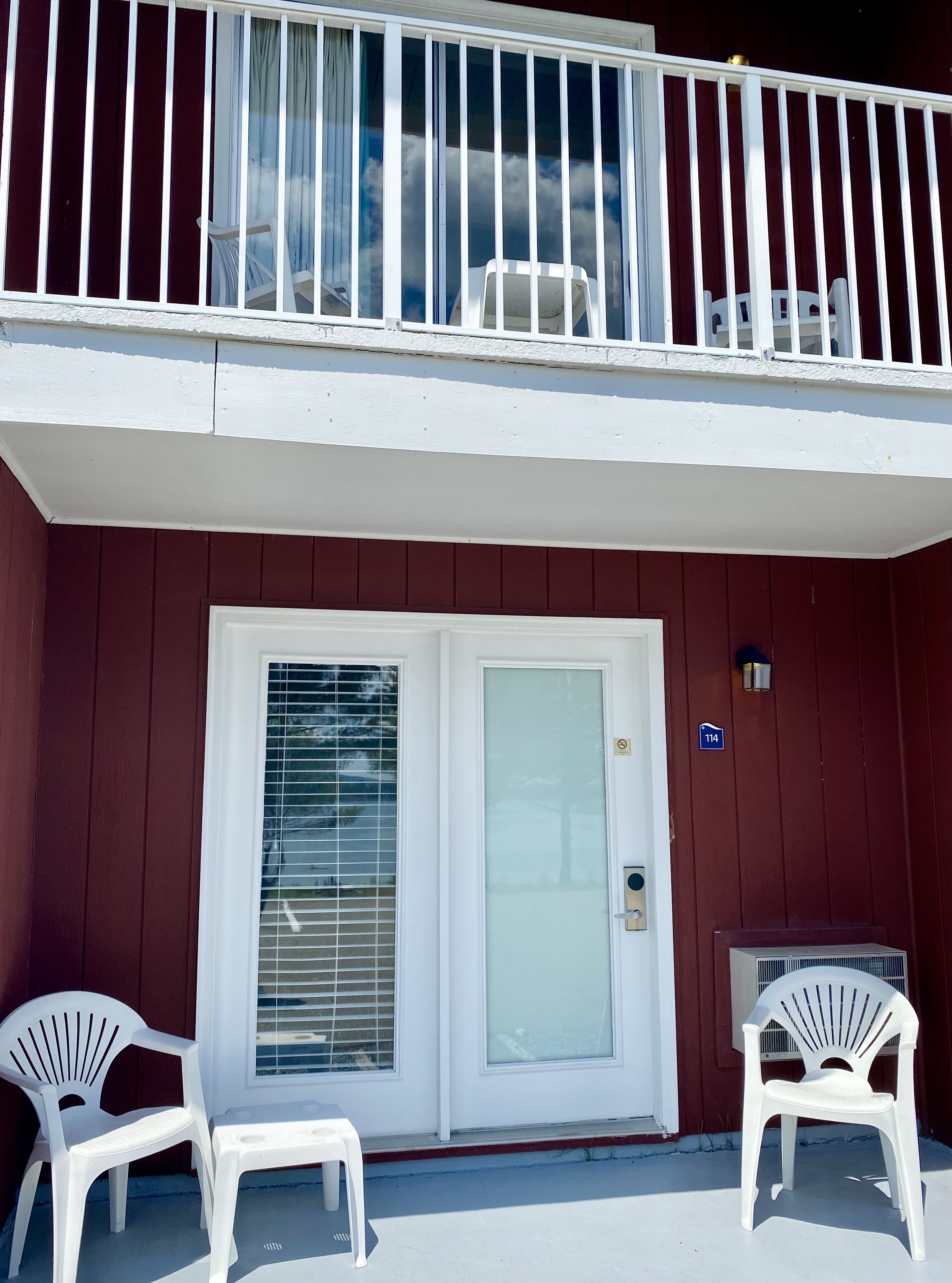 A building with a red exterior, white balcony railing, and a white door with a glass panel. There are two white plastic chairs outside.