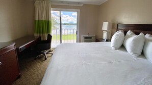 A hotel room with a large bed, a desk, a chair, a nightstand with a lamp, and a view of a body of water through a sliding door. - Island View Hotel Bar Harbor (Trenton)