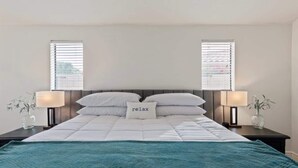 Una cama bien hecha con un edredón color aguamarina, dos lámparas de noche y un cojín con la palabra 'relax'.