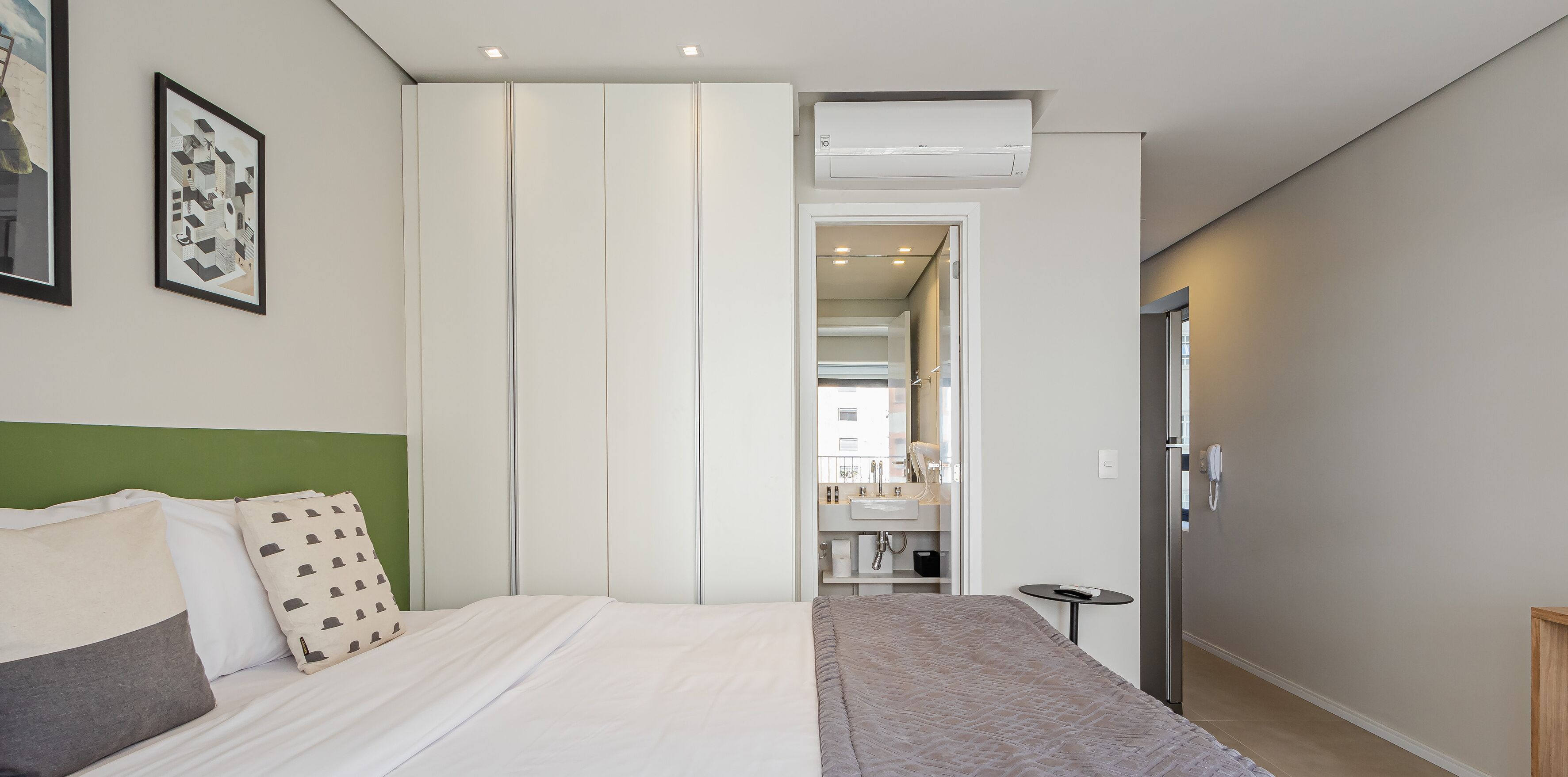 A modern hotel room with a bed, a green headboard, a bedside table, and a bathroom visible through an open door.