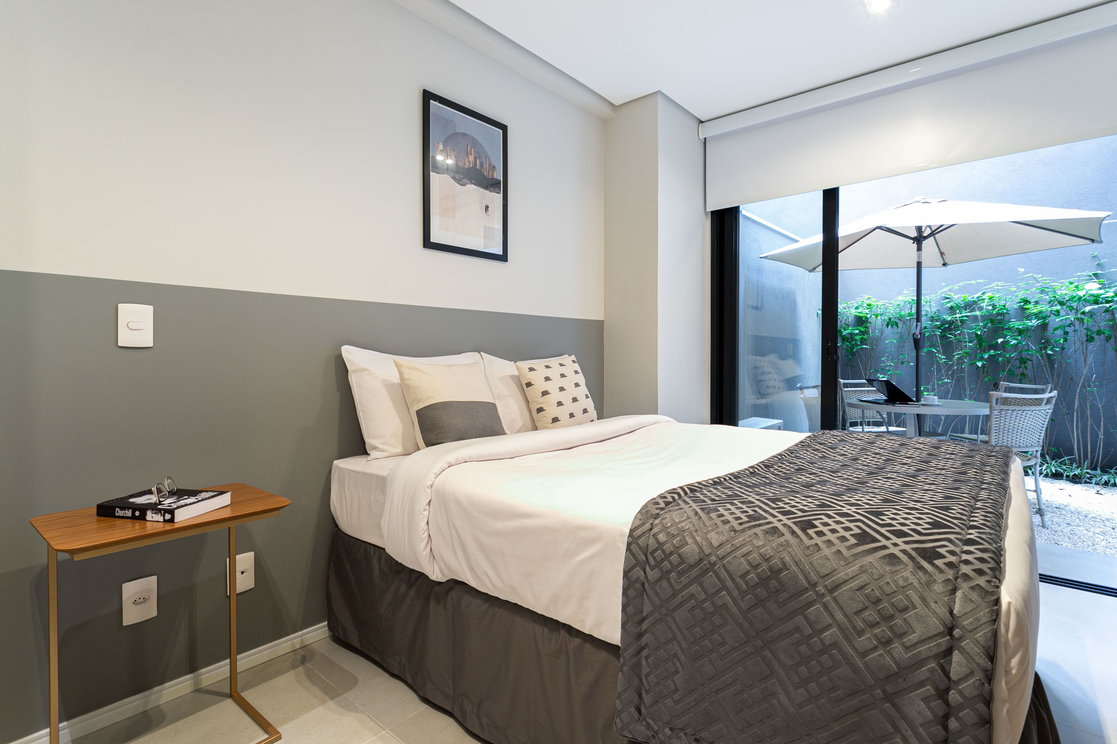 Un dormitorio moderno con cama, mesita de noche y vista a un patio exterior con sombrilla.