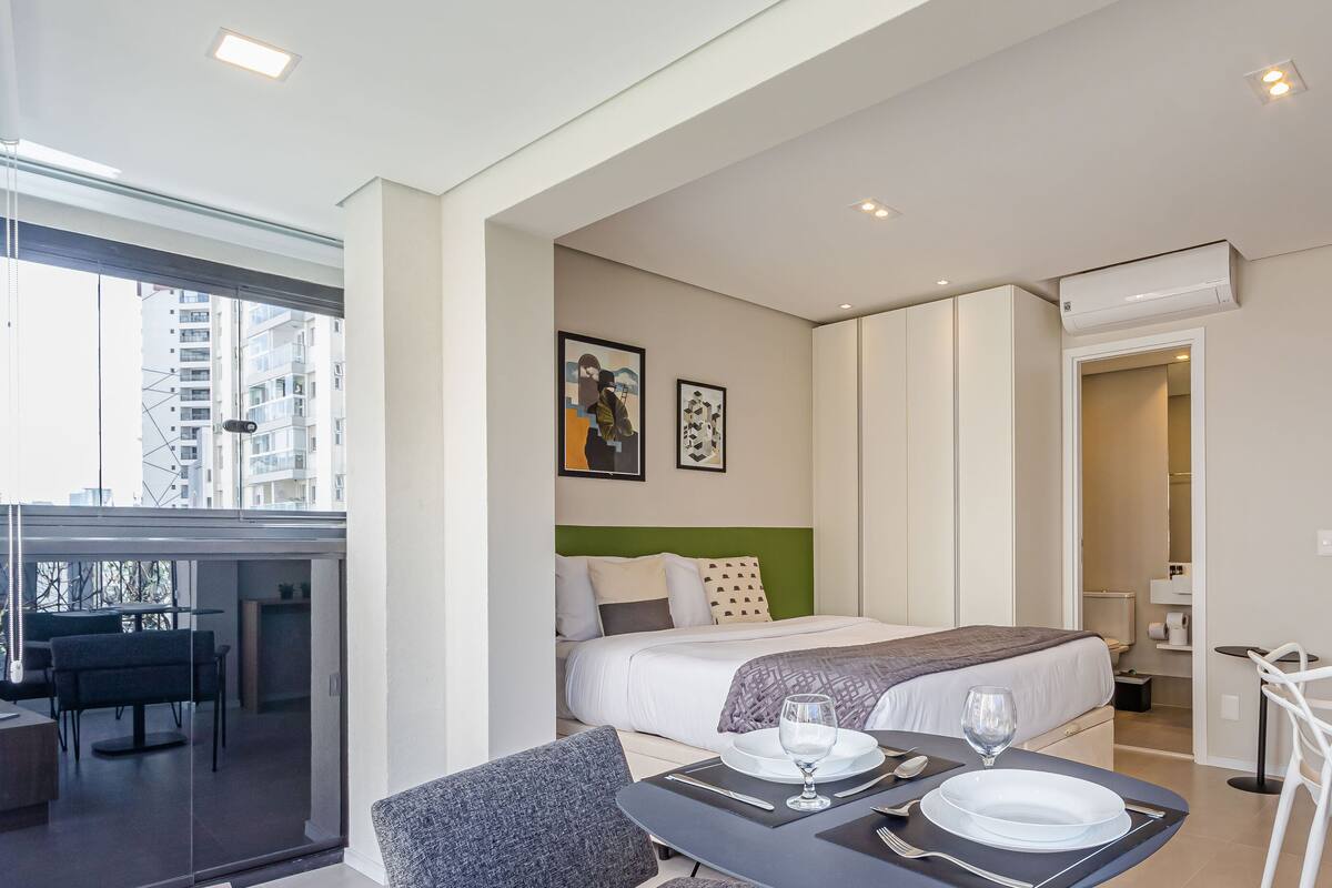 Une chambre d’hôtel moderne, équipée d’un lit, d’une table à manger avec un ordinateur portable, d’une chaise et offrant une vue sur la ville grâce à de grandes fenêtres.