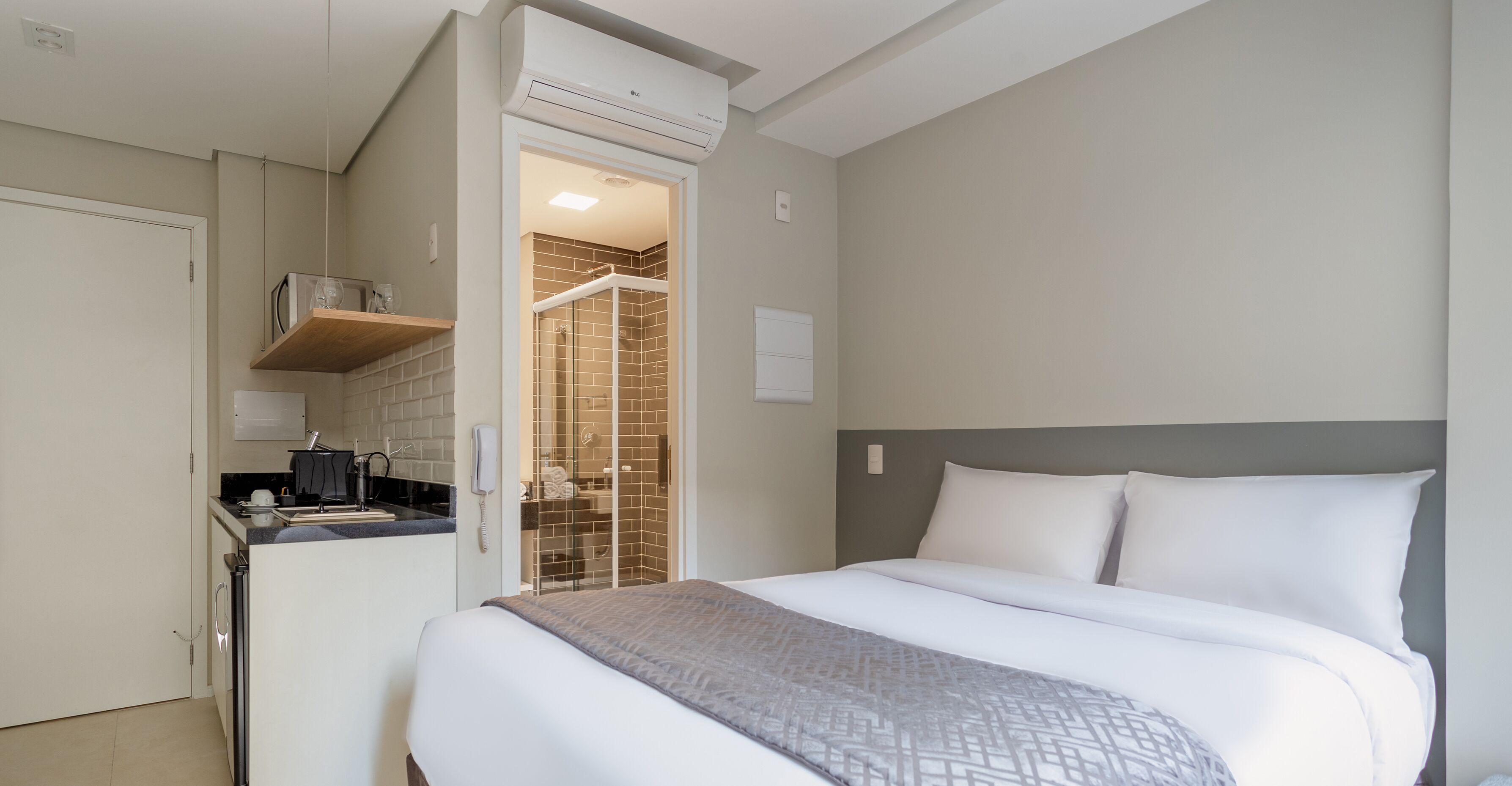 A compact hotel room with a kitchenette, a shower area, and a bed with white bedding.