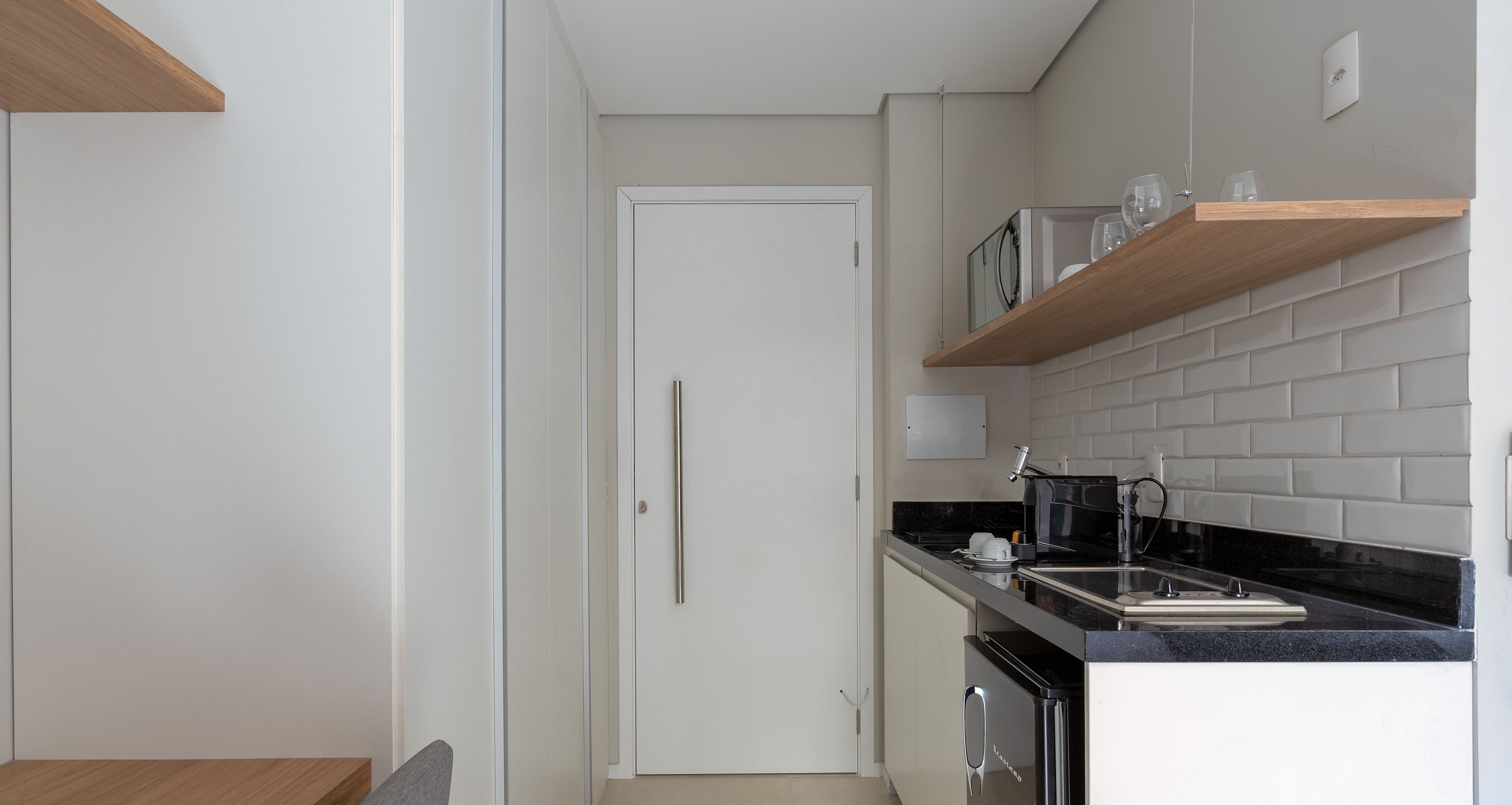 Una cocina compacta con encimera negra, armarios blancos y un estante de madera sobre el lavabo.