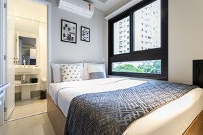 Un dormitorio moderno con una cama grande, una ventana con vistas a edificios y un baño con espejo y lavamanos.