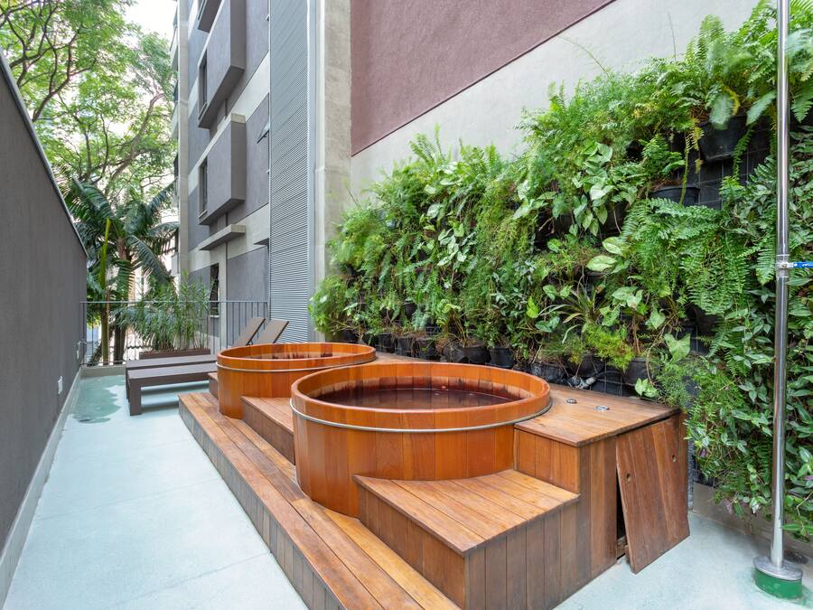 Uma área de estar moderna ao ar livre, com bancos de madeira e floreiras, cercada por vegetação exuberante e fachada de um edifício.