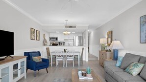 Una sala de estar moderna con televisor, sillón azul, sofá gris, mesa de centro de madera y una zona de comedor con muebles blancos.