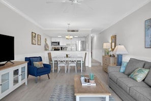 Uma sala de estar moderna com televisão, poltrona azul, sofá cinza, mesa de centro de madeira e uma área de jantar com mobiliário branco.