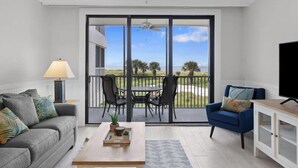 Una sala de estar con sofá, una mesa de centro, televisor, ventilador de techo y un balcón con sillas y una mesa.