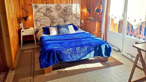 Un dormitorio con pared de paneles de madera, una cama con colcha azul, una mesita de noche, un escritorio y un mapa mundial en la pared.